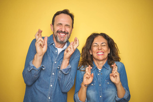 Mooi stel van middelbare leeftijd samen staan over geïsoleerde gele achtergrond gebaren vinger gekruist glimlachen met hoop en ogen dicht. Geluk en bijgelovig concept. - Foto, afbeelding