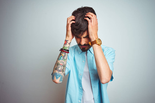 Jovem com tatuagem vestindo camisa azul em pé sobre fundo branco isolado sofrendo de dor de cabeça desesperada e estressada porque dor e enxaqueca. Mãos na cabeça
. - Foto, Imagem
