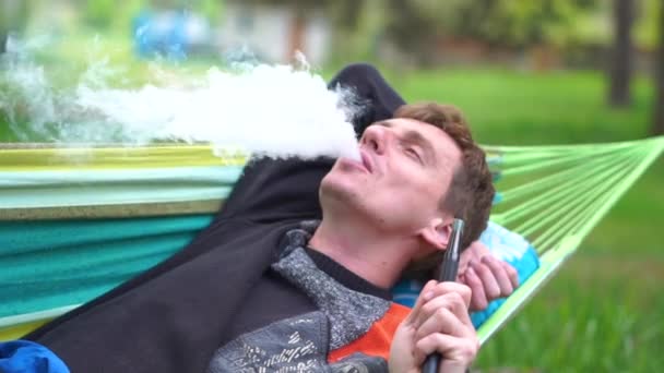 Cheery blond man lying on a wattled hammock smoking hooka in summer in slo-mo - Footage, Video