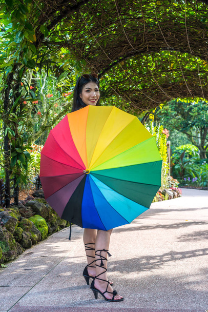 魅力的な美しいアジアの女性女性長い黒髪は、屋外公園の庭でカメラに見て虹のマルチカラー傘を保持します - 写真・画像