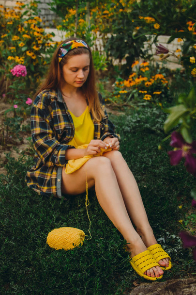 a young girl with long blond hair knits a yellow sweater in the garden in the summer. woman makes clothes with hands closeup - Zdjęcie, obraz