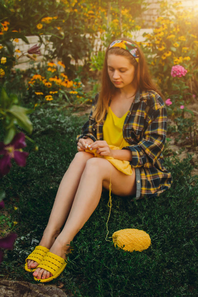a young girl with long blond hair knits a yellow sweater in the garden in the summer. woman makes clothes with hands closeup - Фото, изображение