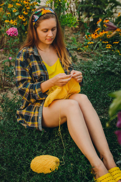 a young girl with long blond hair knits a yellow sweater in the garden in the summer. woman makes clothes with hands closeup - Photo, Image