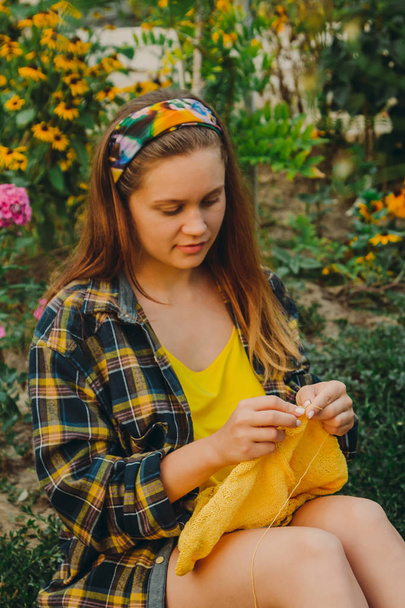 a young girl with long blond hair knits a yellow sweater in the garden in the summer. woman makes clothes with hands closeup - Fotó, kép