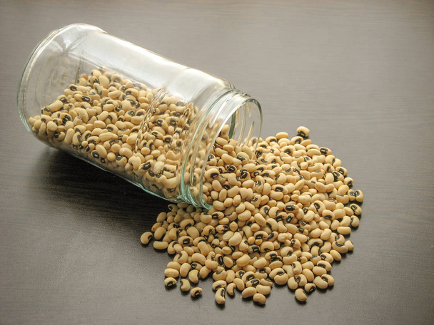 Dried black-eyed peas or beans Vigna unguiculata or cowpea are poured from a glass jar on brown wooden surface.  cowpea in a glass jar on the wooden table.Beans Vigna unguiculata  in a glass dish. Wooden backgroun - Photo, Image