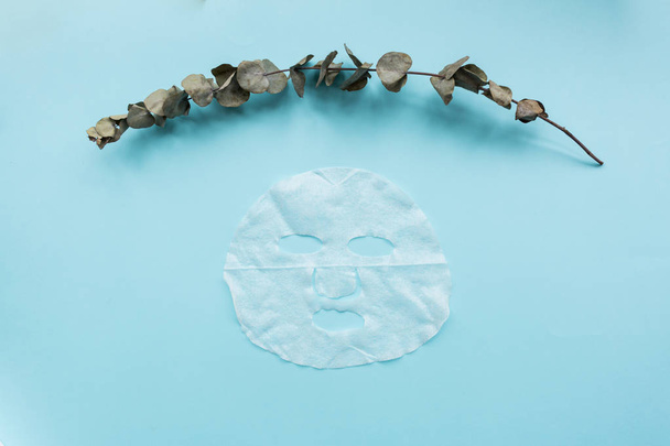 Sheet facial mask and eucalyptus branch on pastel blue paper background. Skin care, dermatology, beauty concept. Top view, flat lay, mockup, overhead, copy space, template - Foto, afbeelding