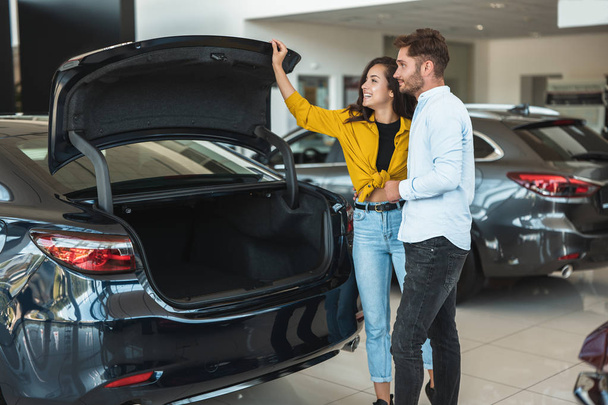 beautiful young couple man and woman looking for a new car in dealership center checking the trunk size - Foto, Bild