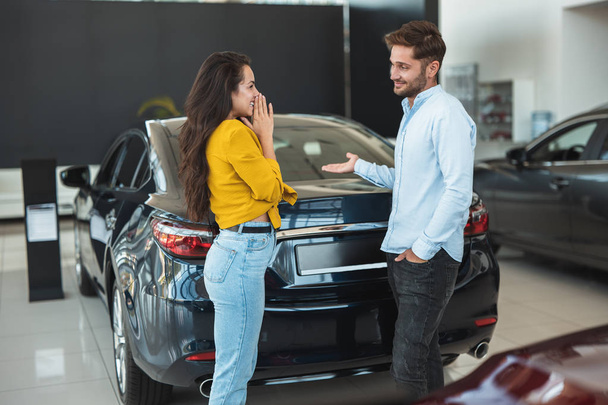 beautiful young couple man and woman deciding which car to choose in dealership center - Zdjęcie, obraz