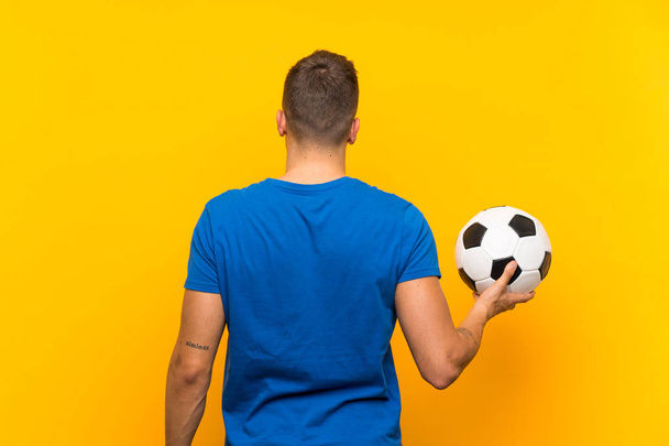 Genç yakışıklı sarışın adam arka konumda izole sarı arka plan üzerinde bir futbol topu tutan - Fotoğraf, Görsel