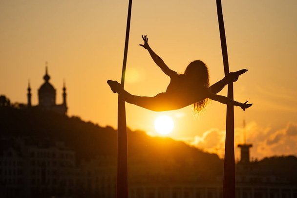 Sylwetka elastycznej kobiety Acrobat na antenę jedwabiu podczas zachodu słońca na tle Kijowa miasta. pojęcie wolności i pokoju - Zdjęcie, obraz