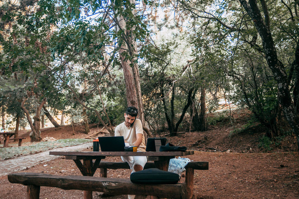 Fiatal szabadúszó vállalkozó online dolgozik egy laptop, miközben ül egy asztalnál a parkban, és beszélni telefonon - Fotó, kép