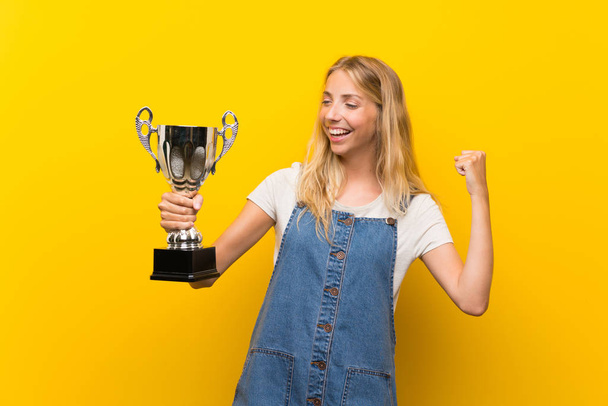 Blonde young woman over isolated yellow background holding a trophy - Photo, Image