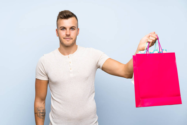 ショッピングバッグの多くを保持する孤立した青い背景の上に若いハンサムなブロンドの男 - 写真・画像