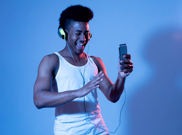 Strong african american young man listening to music online in smartphone app dancing and singing with headphones. Close up portrait with disco dance floor neon light in Music and technology concept. - Foto, Imagen