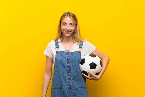 Blonde young woman over isolated yellow background holding a soccer ball - 写真・画像