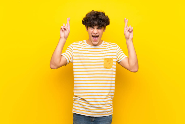Young man over isolated yellow wall with fingers crossing - Fotografie, Obrázek