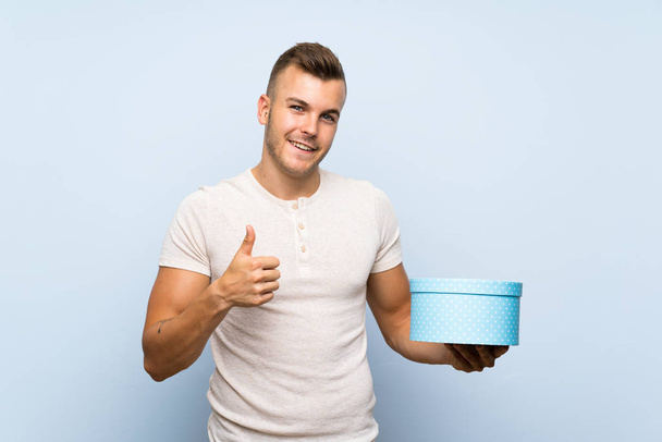 Jonge knappe blonde man over geïsoleerde blauwe achtergrond Holding Gift Box - Foto, afbeelding
