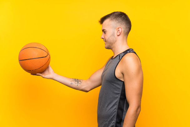 Jeune homme blond beau tenant une balle de basket sur fond jaune isolé avec une expression heureuse
 - Photo, image