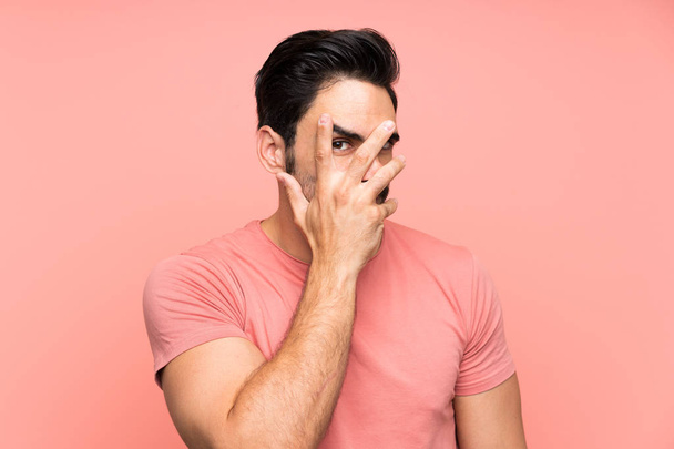Knappe jonge man over geïsoleerde roze achtergrond bedekt ogen en kijken door vingers - Foto, afbeelding