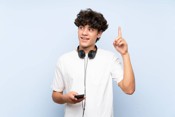junger Mann hört Musik mit einem Handy über isolierte blaue Wand in der Absicht, die Lösung zu realisieren, während er einen Finger hebt - Foto, Bild