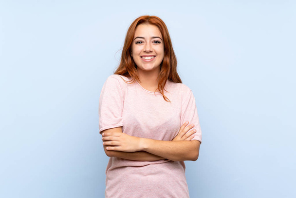 Teenager redhead girl over isolated blue background keeping the arms crossed in frontal position - Fotó, kép