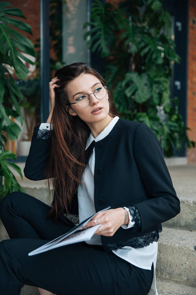 Young beautiful brunette girl, office clothing style, sitting on the street at a table in a cafe, street model posing style, makes notes in a notebook, talking on the phone - Foto, afbeelding
