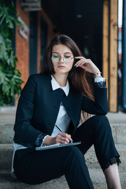 Young beautiful brunette girl, office clothing style, sitting on the street at a table in a cafe, street model posing style, makes notes in a notebook, talking on the phone - Фото, изображение
