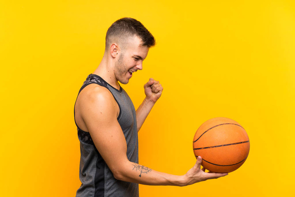 Young handsome blonde man holding a basket ball over isolated yellow background celebrating a victory - Fotografie, Obrázek