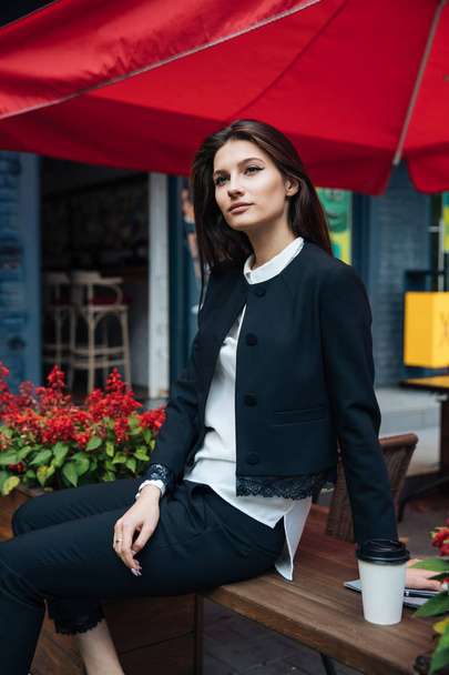 Young beautiful brunette girl, office clothing style, sitting on the street at a table in a cafe, street model posing style, makes notes in a notebook, talking on the phone - 写真・画像
