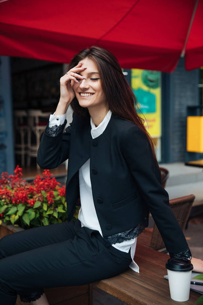Young beautiful brunette girl, office clothing style, sitting on the street at a table in a cafe, street model posing style, makes notes in a notebook, talking on the phone - Zdjęcie, obraz