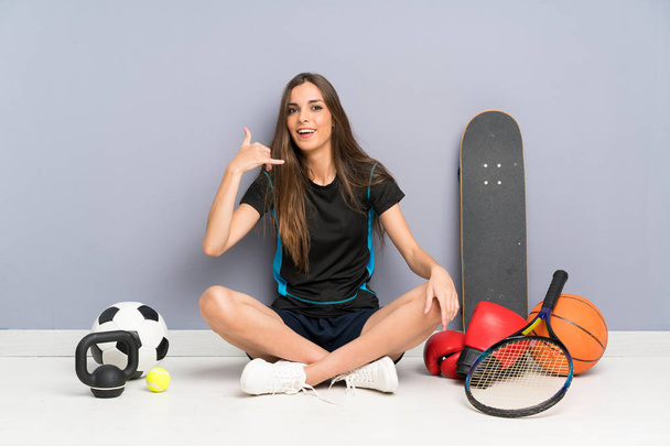 Молода спортивна жінка сидить на підлозі роблячи телефонний жест
 - Фото, зображення