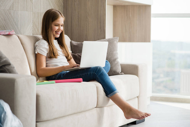 Sorridente ragazza della scuola sedersi con il computer portatile a divano bianco
 - Foto, immagini