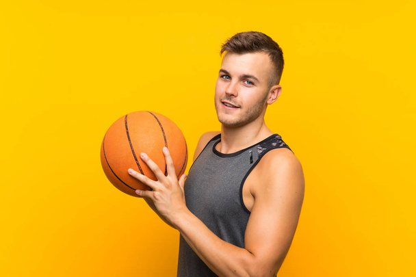 孤立した黄色の背景の上にバスケットボールを保持している若いハンサムなブロンドの男 - 写真・画像