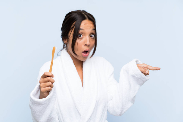 Giovane donna bruna in accappatoio lavarsi i denti su isolato sfondo blu sorpreso e indicando lato
 - Foto, immagini