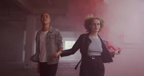Front view of a hip young mixed race man and a hip young Caucasian woman in an empty warehouse, holding hands and running, the woman holding a hand grenade - Filmati, video