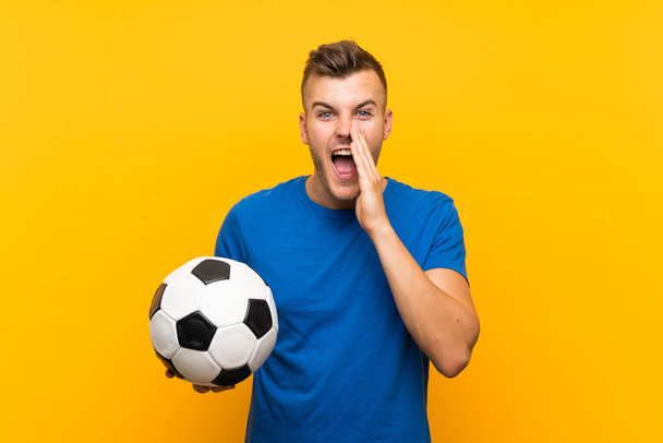 孤立した黄色の背景の上にサッカーボールを持つ若いハンサムなブロンドの男は、口を大きく開いて叫ぶ - 写真・画像