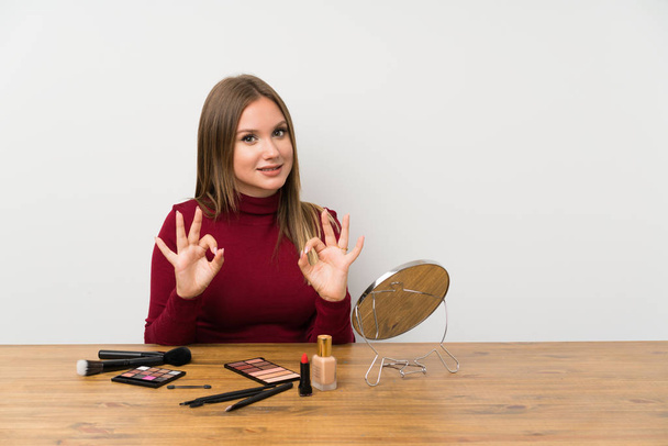 Chica adolescente con paleta de maquillaje y cosméticos en una mesa que muestra un signo de ok con los dedos
 - Foto, Imagen