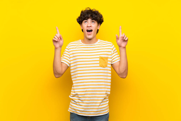 Молодий чоловік над ізольованою жовтою стіною здивований і вказує вгору
 - Фото, зображення
