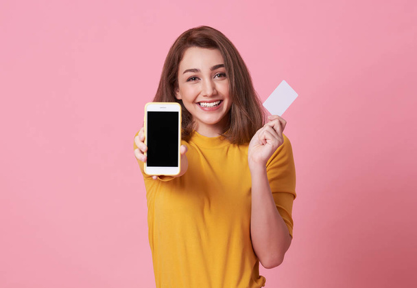 Porträt einer glücklichen jungen Frau, die am leeren Bildschirm Mobiltelefon und Kreditkarte auf rosa Hintergrund zeigt. - Foto, Bild