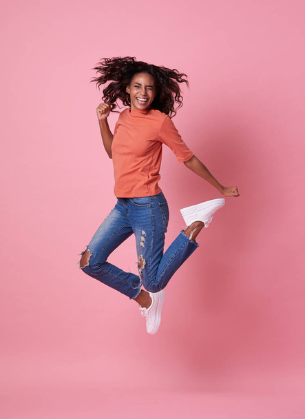 Portrait of a joyful young african woman in orange shirt jumping and celebrating over pink background. - Photo, Image