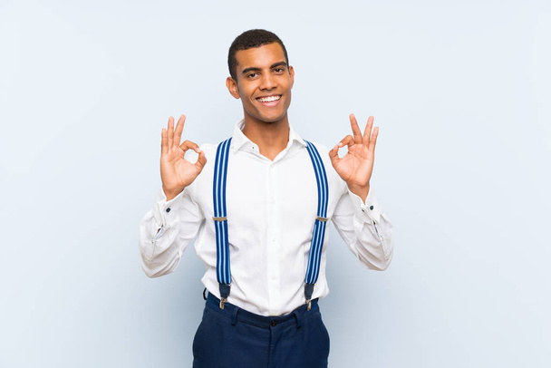 Mladý pohledný brunetý muž s kšandama na izolovaném pozadí ukazující dobré znamení s prsty - Fotografie, Obrázek