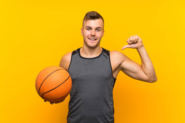 Jonge knappe blonde man houdt een basket ball over geïsoleerde gele achtergrond trots en zelf-tevreden - Foto, afbeelding