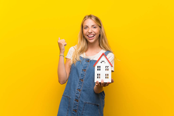 Блондинка молода жінка над ізольованим жовтим тлом тримає маленький будинок
 - Фото, зображення