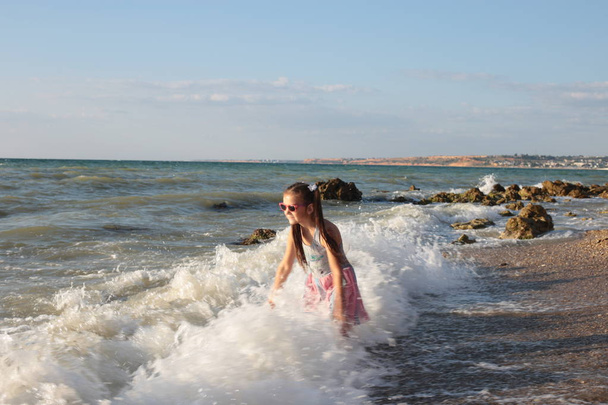 Pieni ilkikurinen ja iloinen kahdeksan vuoden tyttö, jolla on pitkät ruskeat hiukset hyppää aaltoihin ja nauraa kesälomien ilolla.
 - Valokuva, kuva