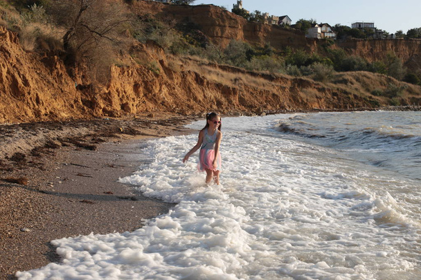 ein kleines schelmisches und fröhliches Mädchen von acht Jahren mit langen braunen Haaren springt auf die Wellen und lacht vor Freude über die Sommerferien - Foto, Bild