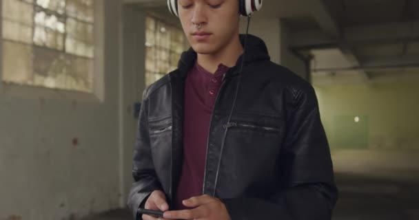 Front view of a hip young mixed race man in an empty warehouse, holding a smartphone listening to music with headphones on and eyes closed - Πλάνα, βίντεο
