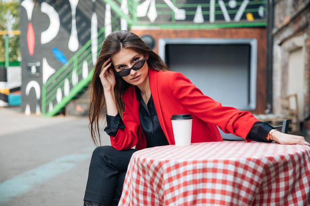 Young beautiful brunette girl, office clothing style, sitting on the street at a table in a cafe, street model posing style, makes notes in a notebook, talking on the phone - Foto, imagen