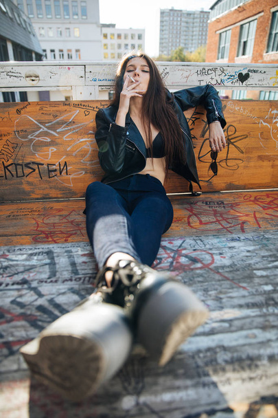 deri ceketli genç kız sigara içiyor, rock tarzı, arsız ve sevimli kız sokak poz tarzı, rock punk yaşam tarzı - Fotoğraf, Görsel