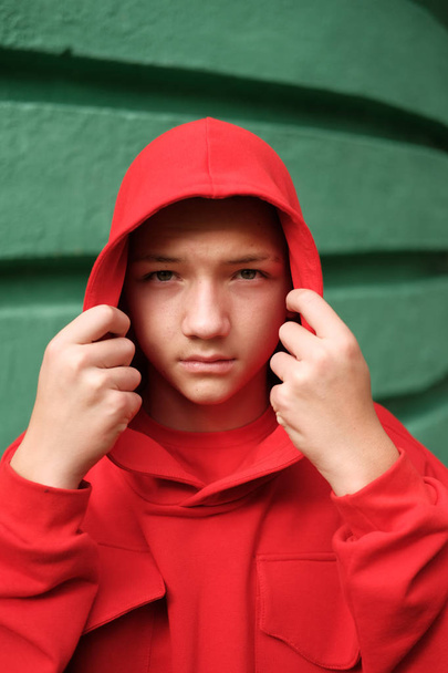 stylish casual teenage boy in fashionable red hoodie against green wall - Фото, изображение