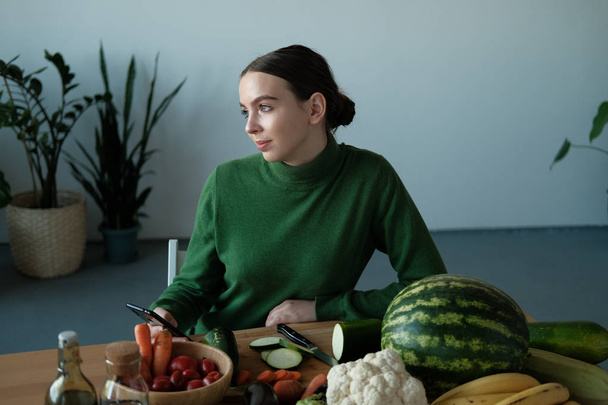 young vegan woman using phone at kitchen table with veggies in home interior - Zdjęcie, obraz